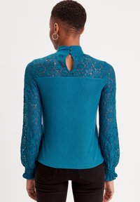Top a maniche lunghe color teal con dettagli in pizzo sulle maniche, dotato di chiusura a bottone a oblò sul retro e corpino testurizzato.