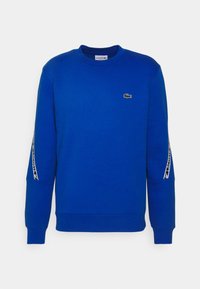 Modrý svetřík vyrobený z bavlny, s kulatým výstřihem a dlouhými rukávy. Logo Lacoste na hrudi, bílé označení podél rukávů.