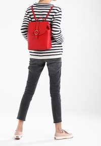 Zaino rosso in ecopelle con parte superiore arrotolata e accessori circolari, indossato sulla schiena. Maglione a righe e pantaloni grigi visibili.