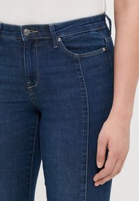 Tamno plave traperice s glatkom teksturom, koje imaju klasičan dizajn s pet džepova, metalnu kopču i suptilne detalje šivanja.