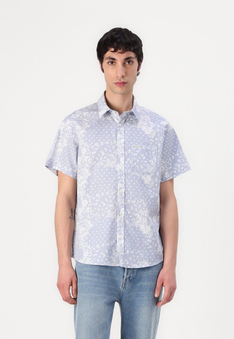 Camicia a maniche corte con colletto in tessuto azzurro chiaro, decorata con motivi floreali e a pois, dotata di taschino sul petto e chiusura con bottoni.