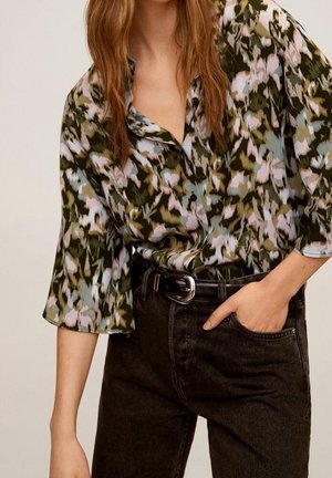 Person iført en løs blomstret knap-up bluse i pastelfarver og mørke højtaljede jeans med sort bælte, hånd i lommen.