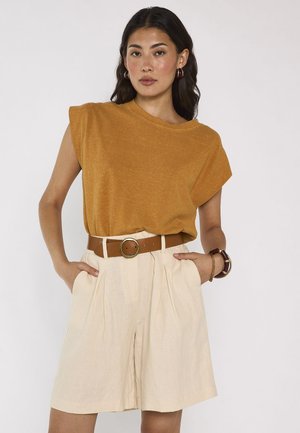 Femme portant un haut sans manches moutarde, un short beige taille haute à plis, une ceinture marron, des boucles d’oreilles créoles et des bracelets, debout avec assurance.