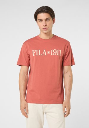 Młody mężczyzna o krótkich, ciemnych włosach, ubrany w rdzawobrązową koszulkę z logo FILA 1911 oraz jasnobeżowe spodnie, stojący na jednolitym tle.