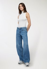 Top a maniche senza e colletto a imbuto bianco abbinato a jeans blu a vita alta e a gamba larga. I jeans presentano un classico design a cinque tasche e una finitura sbiadita.