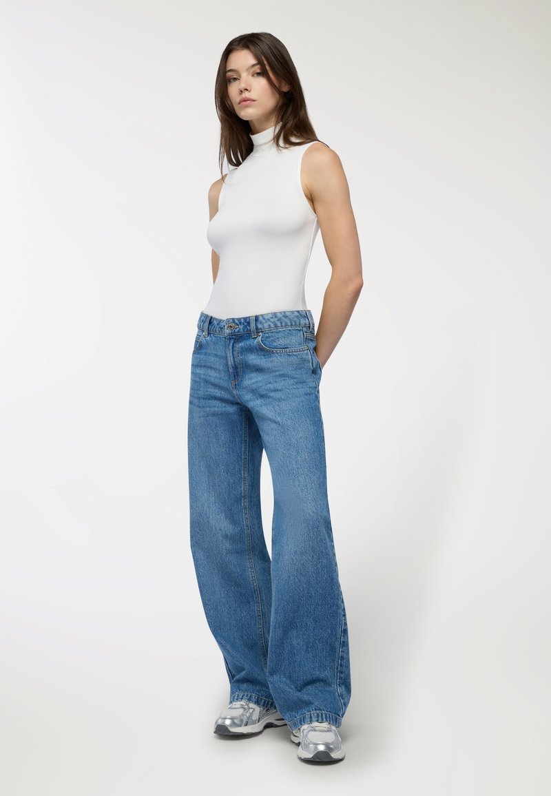 Top a maniche senza e colletto a imbuto bianco abbinato a jeans blu a vita alta e a gamba larga. I jeans presentano un classico design a cinque tasche e una finitura sbiadita.