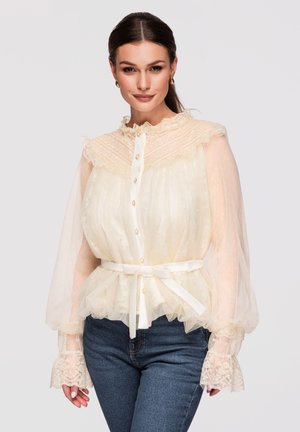 Mujer con blusa de encaje crema transparente con cuello con volantes, mangas globo, botones de perla y una cinta en la cintura, combinada con vaqueros azules.