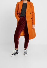 Manteau croisé en mélange de laine orange, associé à un pantalon en velours côtelé bordeaux et des baskets montantes noires. Texture lisse, coupe ajustée.