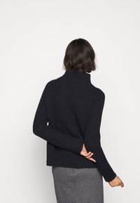 Pull à col roulé noir à côtes avec des manches ajustées, présentant un design en tricot texturé et une coupe décontractée à l'ourlet, associé à une jupe à motifs.