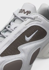 Λευκό και γκρι αθλητικό παπούτσι Nike με δίχτυ στο πλάι, γκρι λογότυπο Nike swoosh, λευκά κορδόνια και το λογότυπο «AMP» στη γλώσσα.