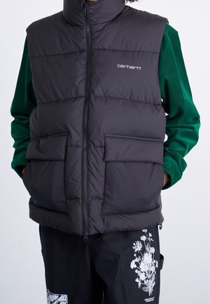 Personne portant une doudoune sans manches noire Carhartt sur une chemise verte à manches longues et un pantalon noir à imprimé floral blanc.