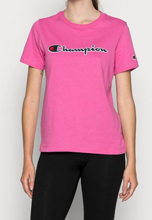 Femme portant un t-shirt Champion rose vif avec le logo sur la poitrine et sur la manche gauche, associée à un pantalon noir, debout avec les bras le long du corps.