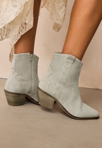 Lichtgrijze suède enkellaarsjes met een spitse neus, een gestapelde hak en decoratieve stiksels, voorzien van een zijrits als sluiting.