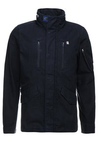 Mörk marinblå jacka i bomull med ståkrage, dragkedjefickor på bröstet och sidofickor med lappar. Har en logotyp på vänster bröst.