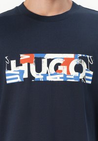 Mørkeblå bomulls T-skjorte med et slitt grafisk logo "HUGO" i hvit, rød og blå, sentrert over brystet. Korte ermer, rund hals.