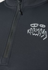 T-shirt de sport noir avec fermeture éclair, présentant un panneau avant texturé et un design graphique blanc avec des motifs de mains et le mot "KARMA."