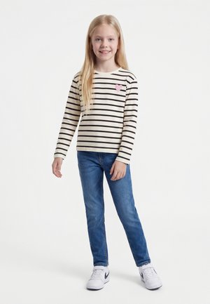 Noor tüdruk pika blondide juustega, kandes valget ja musta triibulist pika varrukaga särki, millel on roosa süda, siniseid teksaseid ja valgeid tenniseid.