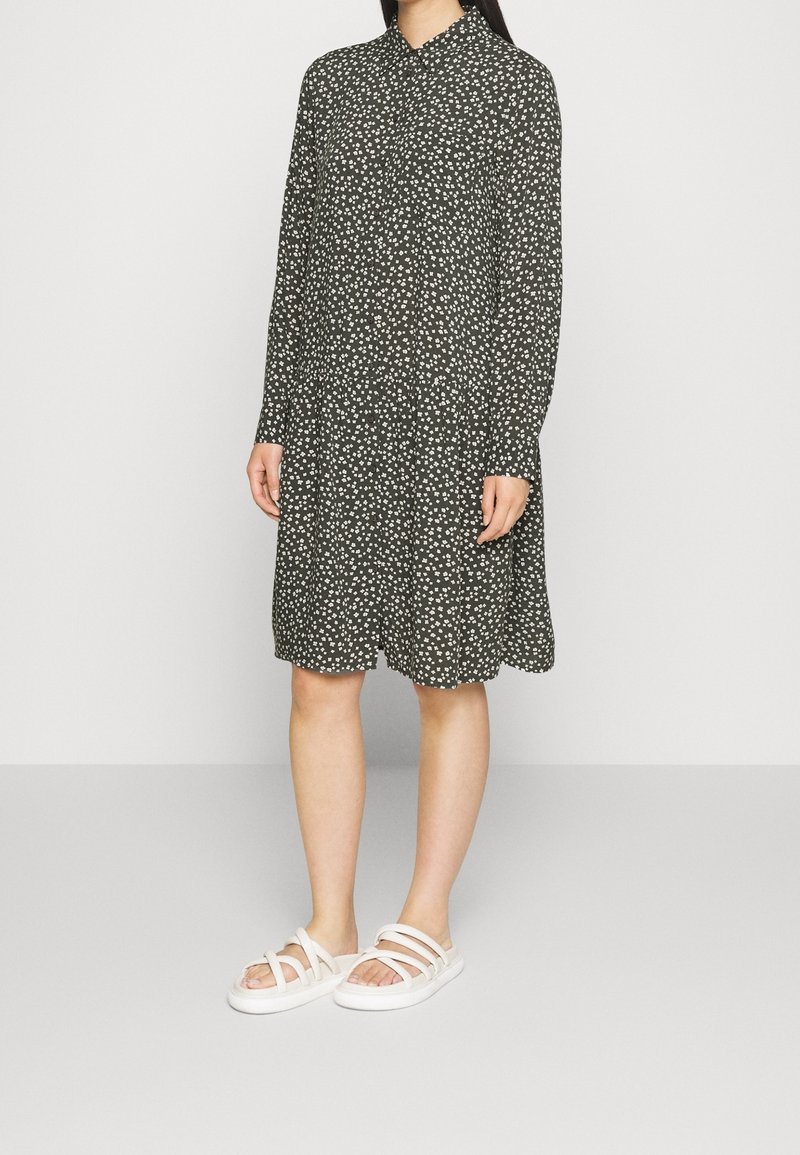 Robe noire avec motif floral blanc, manches longues, design col, longueur genoux, associée à des sandales blanches à lanières sur un fond neutre.