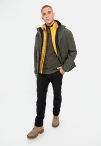 Herre lagdelte outfit med en olivengrøn jakke, gul quiltet vest, grå sweater, sorte bukser og beige støvler.