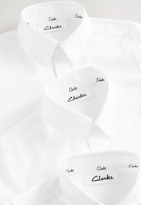 Zwei weiße Hemden übereinander, mit klassischen Kragen. Jeder Kragen zeigt den Markennamen "Clarks" in schwarzem Stich entlang des Bandes.