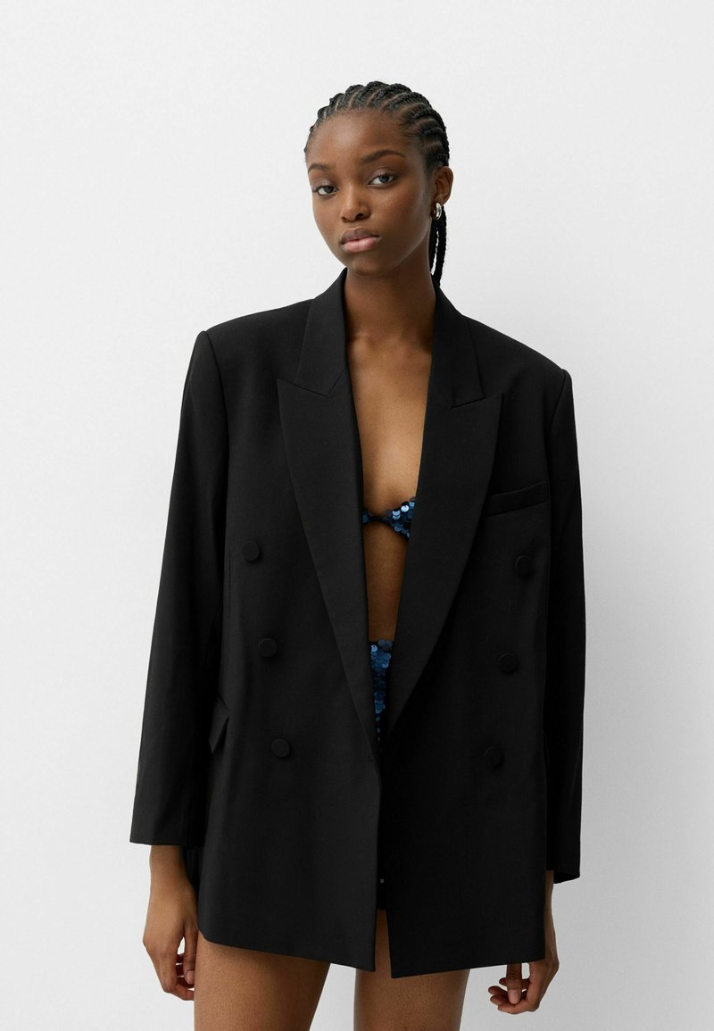 Blazer noir à double boutonnage avec une coupe décontractée, présentant des revers crantés, de grands boutons et des poches avant. Tissu lisse et design ajusté.