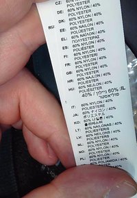 Etiqueta de ropa que muestra la composición del tejido: 60% nylon y 40% poliéster, en varios idiomas, sostenida por una mano.