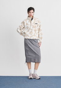 Γυναίκα που στέκεται, φοράει μπεζ και λευκή φλις μπλούζα με σχέδιο, γκρι μίντι φούστα με τσέπες, λευκές κάλτσες και ροζ αθλητικά παπούτσια σε μπλε δάπεδο.