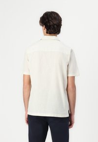 Camicia a maniche corte in tessuto beige chiaro con finitura texturizzata, dotata di colletto a punta e orlo dritto. Nessun logo o motivo visibile.