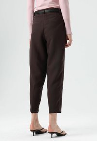 Bruine, op maat gemaakte broek met taps toelopende pijpen, voorzien van een vlakke voorkant, zijzakken en een slanke zwarte riem, die boven de enkel valt.