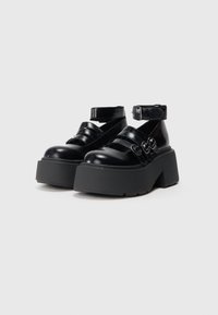 Zwarte platform schoenen van glanzend materiaal met drie banden en ronde gespen. Voorzien van een dikke rubberen zool en enkelband.