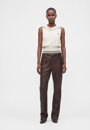 Femme aux cheveux courts portant un haut blanc sans manches avec col et un pantalon en cuir marron foncé taille haute sur fond blanc.