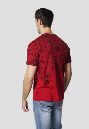Camiseta roja de algodón con mangas cortas, con un patrón abstracto negro y un logo cerca del dobladillo. Combinada con vaqueros de color azul claro.