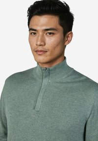 Grüner Pullover mit Reißverschluss und hohem Kragen, hergestellt aus weichem Strickstoff. Verfügt über eine feine Textur und einen kurzen Reißverschluss am Hals.