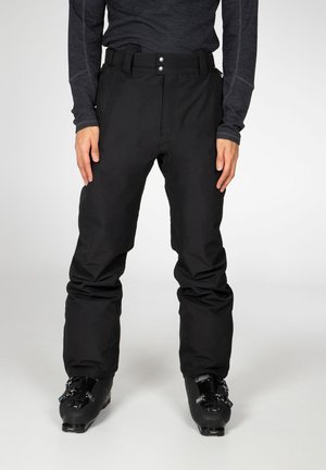 Person trägt schwarze Skihose und schwarze Skischuhe und steht vor einem schlichten Hintergrund.