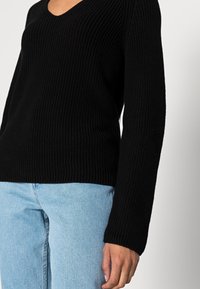 El suéter de punto acanalado negro presenta un escote en V y mangas largas, combinado con denim azul claro. La textura es gruesa y acogedora.
