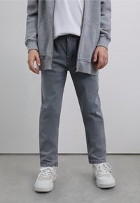 Sweat à capuche gris zippé sur un t-shirt blanc, associé à un jean gris clair et des baskets blanches. Les matériaux semblent doux et décontractés.