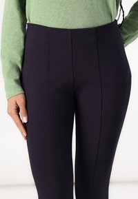 Kvinna som bär högmidjade mörkblå tighta byxor och en ljusgr ön långärmad topp, stående med ena handen vilande på höften.