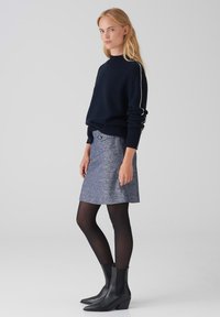 Marineblauwe gebreide trui met een geribbelde halslijn, gecombineerd met een gestructureerde blauwe rok met knoopdetail, zwarte panty’s en puntige enkelboots.