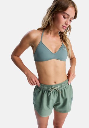 Femme ajustant un short de bain vert avec taille élastique, portant un haut de bikini assorti vert, sur fond blanc uni.