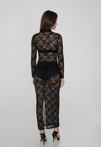 Vestido de encaje negro con mangas largas, tela semi-translúcida, cuello alto y aberturas hasta el muslo, combinado con una prenda interior negra.