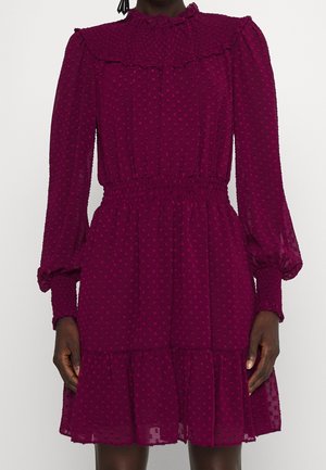 Kobieta ubrana w teksturowaną sukienkę wiśniowego koloru za kolano, z długimi rękawami, marszczoną talią, falbankowym dekoltem i mankietami na rękawach.