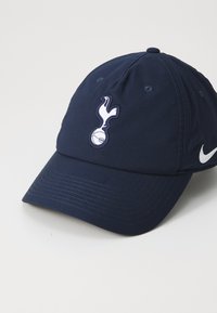 Marinblå baseballkeps med broderad Spurs-logotyp i vitt, tillverkad av textilmaterial. Har en böjd skärm och Nike-logotyp i vitt.