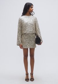 Σετ με τοπ και μίνι φούστα διακοσμημένα με λαμπερά χρυσά παγιέτες· μακριά μανίκια· άνετη εφαρμογή· συνδυάζεται με μαύρη τσάντα χειρός· μοντέλο σε πέδιλα με λουράκια και ανοιχτά δάχτυλα.