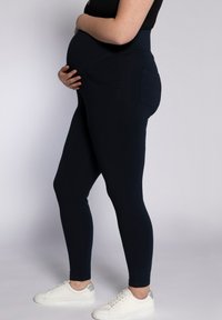 Leggings premaman di colore blu navy scuro, in tessuto elasticizzato con vita alta e tasche laterali, abbinati a sneaker bianche su uno sfondo semplice.