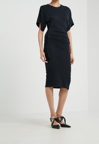 Svart figurnära klänning med korta ärmar, samlad sidodetalj, knälång fåll, i kombination med svarta spetsiga klackskor med dekorativa remmar.