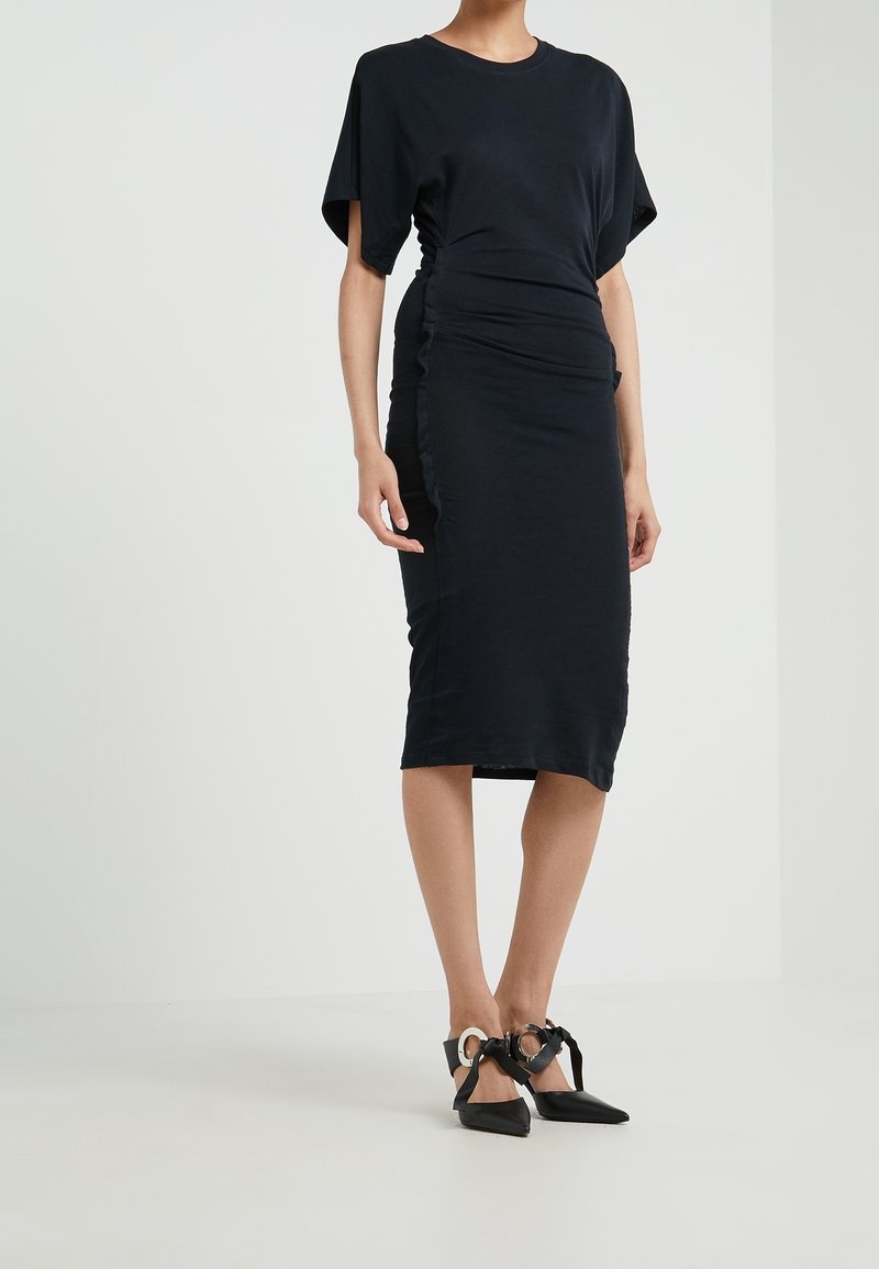 Svart figurnära klänning med korta ärmar, samlad sidodetalj, knälång fåll, i kombination med svarta spetsiga klackskor med dekorativa remmar.