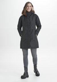 Cappotto impermeabile nero con cappuccio, chiusura con zip e regolazioni a cordoncino. Presenta tasche laterali e un design fino al ginocchio. Indossato con jeans grigi e stivali neri.