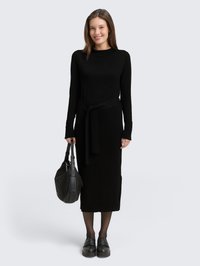 Robe noire à manches longues avec un détail de ceinture à nouer, fabriquée en tissu doux. Présente une silhouette droite et est portée avec des chaussures à plateforme noires et un sac.