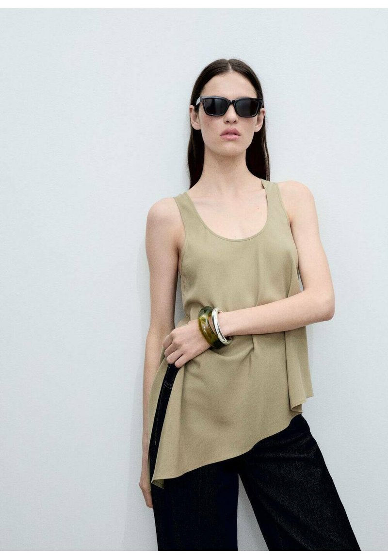 Khaki tanktop met een gedrapeerd ontwerp, gedragen met donkere jeans. Accessoires omvatten meerkleurige armbanden om één pols en slanke zwarte zonnebril.