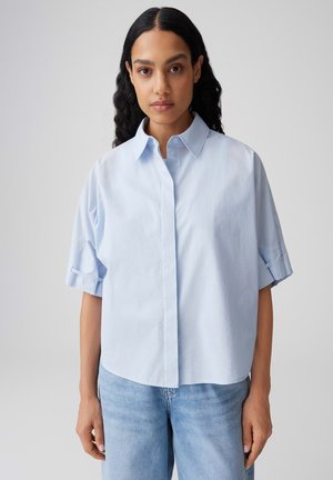 Femme aux longs cheveux châtains portant une chemise oversize à manches courtes bleu clair et un jean bleu clair, debout devant un fond uni.